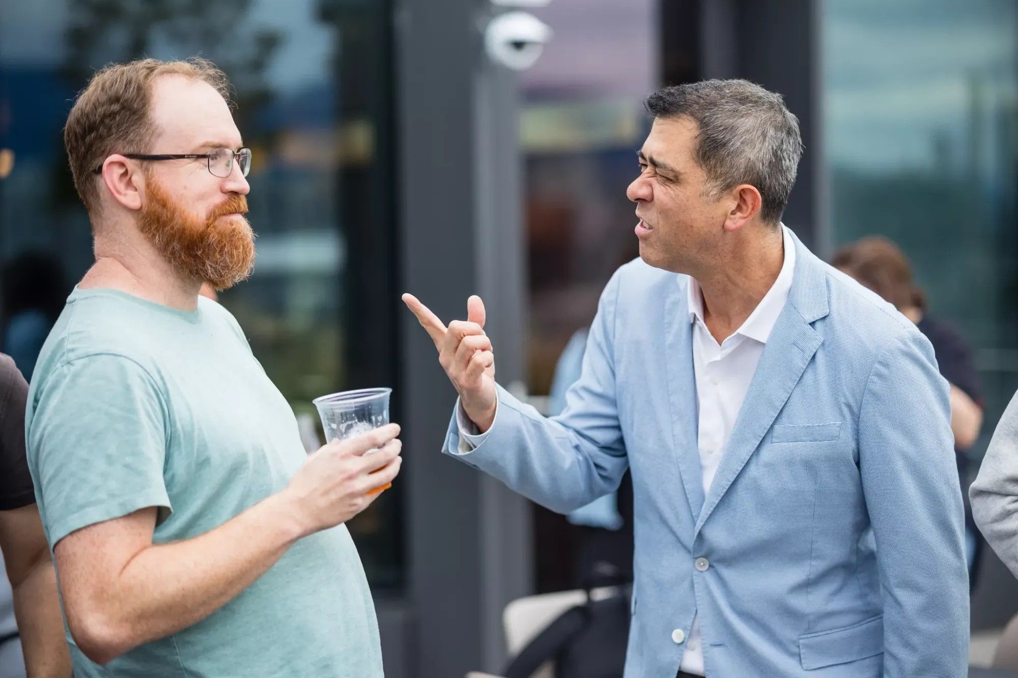 Darin Grant in a light blue blazer, mid-sentence and pointing a finger at a bearded colleague holding a drink, at an outdoor gathering