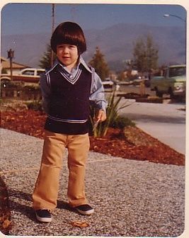 Darin Grant as a small child in a sweater vest and flared trousers, standing on a driveway with hills behind