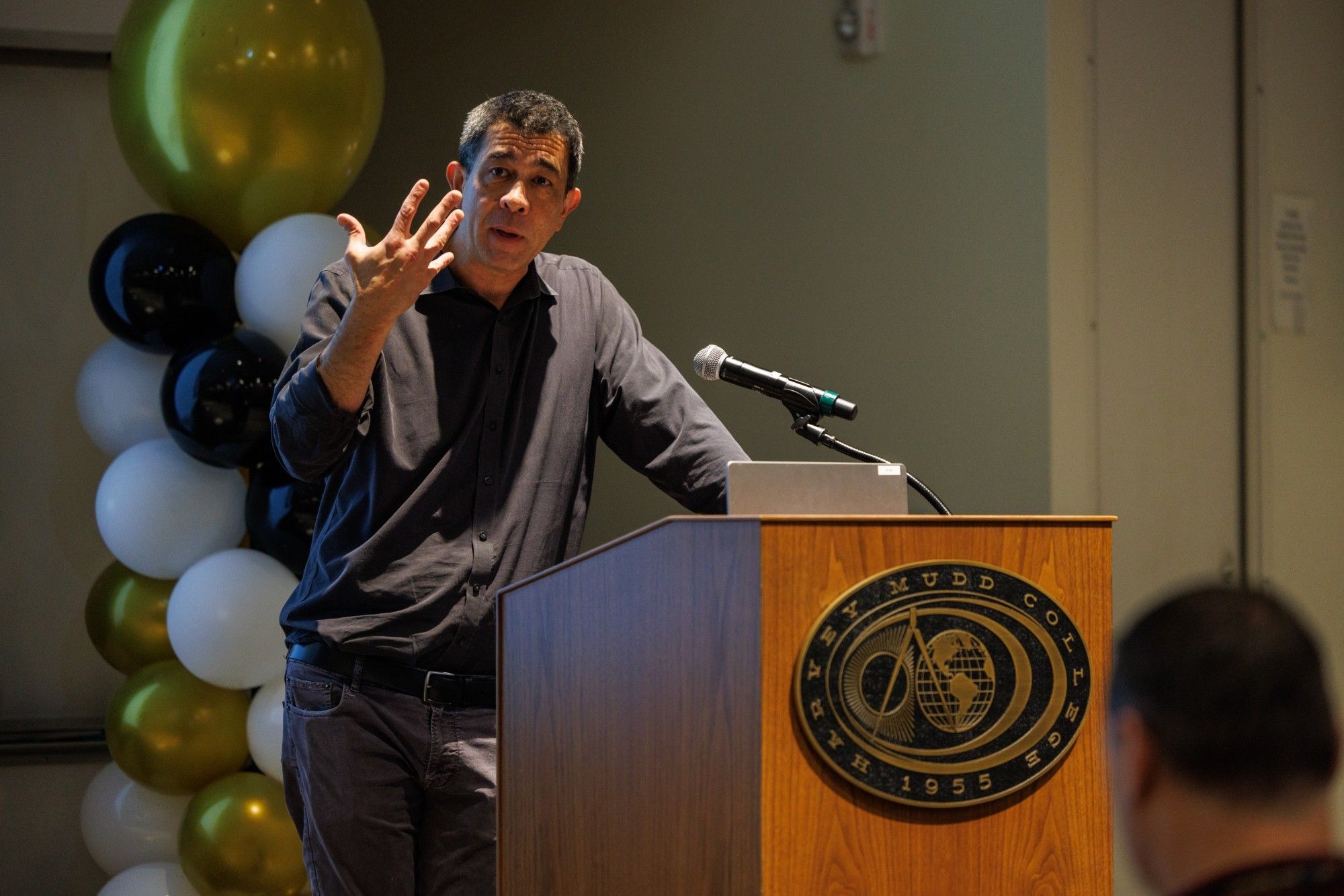 Darin Grant speaking and gesturing at a Harvey Mudd College podium, with balloons behind him